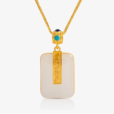 A 24K Gold and Nephrite Pendant with a rectangular nephrite stone adorned by 24K gold detailing and an engraved six-syllable pattern. The bail is accented with turquoise and sapphire stones for added elegance.